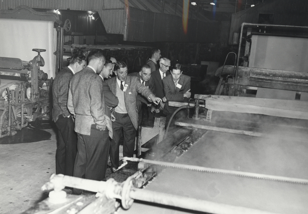 Photographie noir et blanc représentant un groupe d’hommes se tenant près d’une machine à papier. En arrière-plan, on aperçoit une autre machine, à gauche, et l’intérieur de l’usine. 