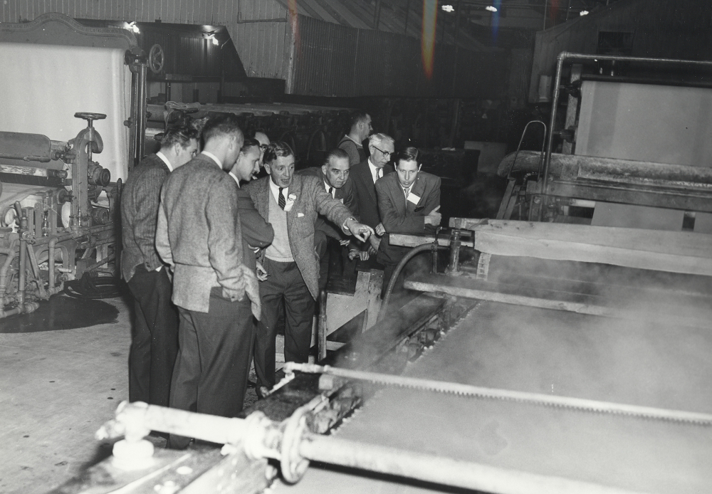 Photographie noir et blanc représentant un groupe d’hommes se tenant près d’une machine à papier. En arrière-plan, on aperçoit une autre machine, à gauche, et l’intérieur de l’usine.