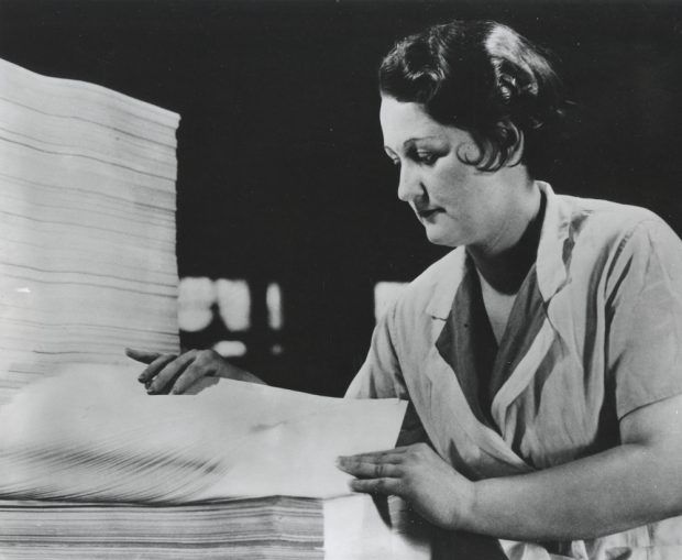 Photographie en noir et blanc représentant une femme en train de compter une pile de feuilles. 