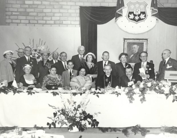Photographie noir et blanc d’un groupe de personnes à une table d’honneur lors d’une célébration. 