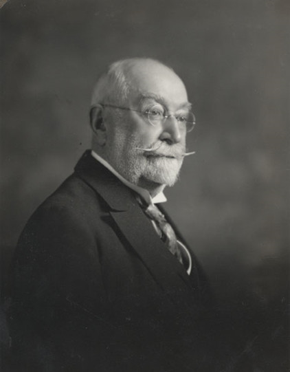 Photographie noir et blanc d’un homme âgé, ayant une courte barbe blanche et une longue moustache retroussée. Il porte des lunettes rondes, une chemise blanche avec une cravate ainsi qu’un veston noir. 
