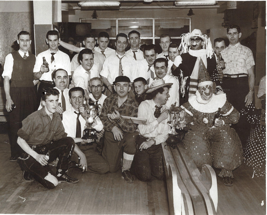 Photographie noir et blanc d’un groupe d’hommes, certains costumés, près d’une allée de quilles. 