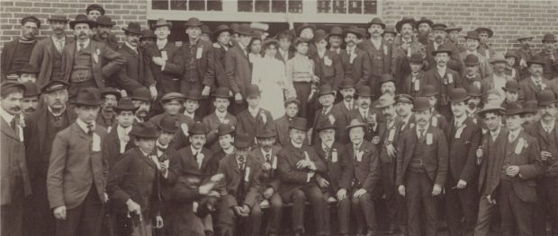 Photographie sépia illustrant des hommes et quelques femmes à l’extérieur devant une usine. 