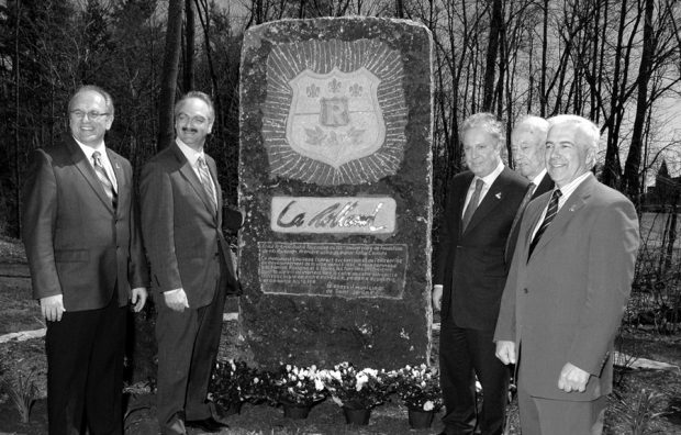 Photographie noir et blanc de cinq hommes près d’un monument en granite à l’extérieur. 
