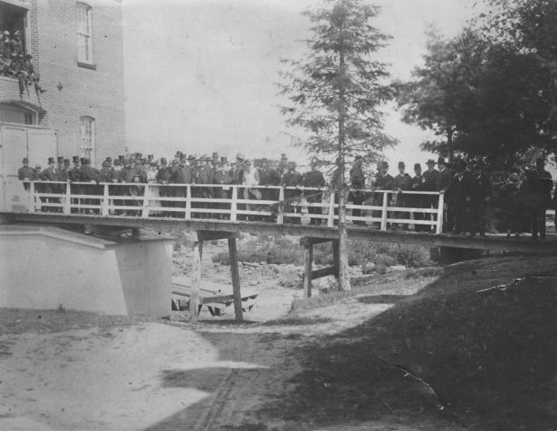 Photographie noir et blanc représentant plusieurs personnes, tant des hommes que des femmes, debout sur un pont. On aperçoit une partie de l’usine de Saint-Jérôme à gauche ainsi que plusieurs arbres à droite. 