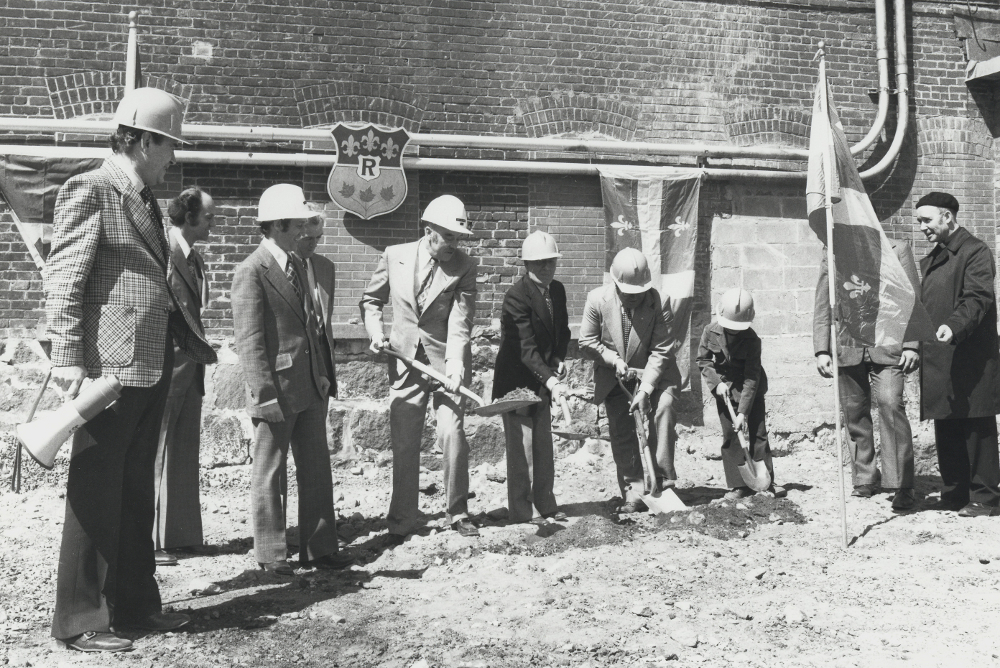 Photographie noir et blanc représentant plusieurs hommes debout un à côté de l’autre, habillés de complets et portant des casques de construction, en train de pelleter. 