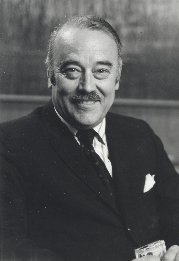 Portrait noir et blanc d’un homme souriant dans la quarantaine ou la cinquantaine. Il porte une petite moustache ainsi qu’un complet veston cravate noir. 