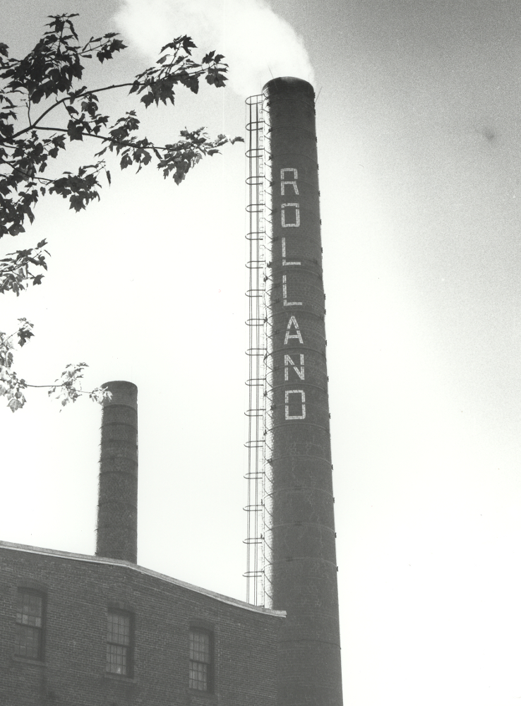 Photographie noir et blanc d’une cheminée d’une usine sur laquelle est inscrit le nom Rolland. 
