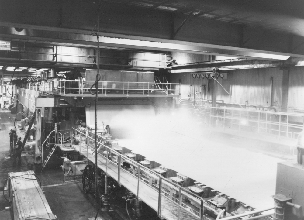 Photographie noir et blanc d’une machine à papier en fonctionnement. De la vapeur s’échappe d’un des plateaux, visible en avant-plan. À gauche, on aperçoit des ouvriers près de la machine. 