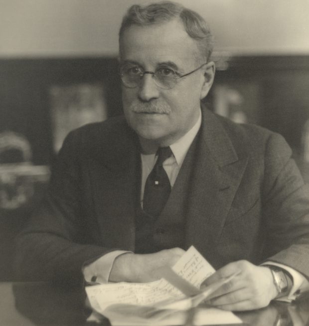 Portrait noir et blanc d’un homme, portant des lunettes et un complet, assis à un bureau, une feuille de papier à la main. 