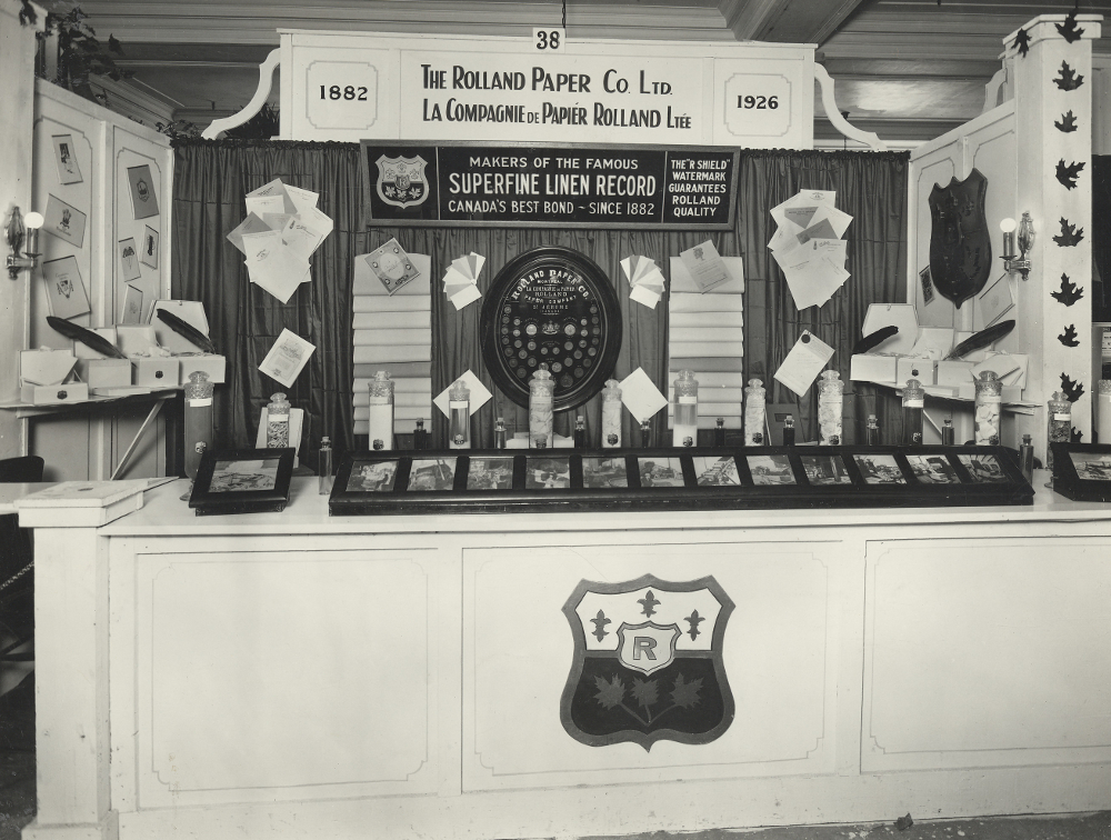 Photographie noir et blanc d’un kiosque où sont présentés plusieurs photographies, échantillons de papier et contenants en verre. Des affiches annoncent le nom de la Compagnie Rolland. 