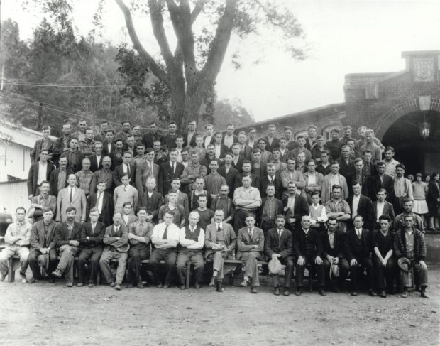 Photographie noir et blanc d’un grand groupe d’hommes en rangées devant une usine.  