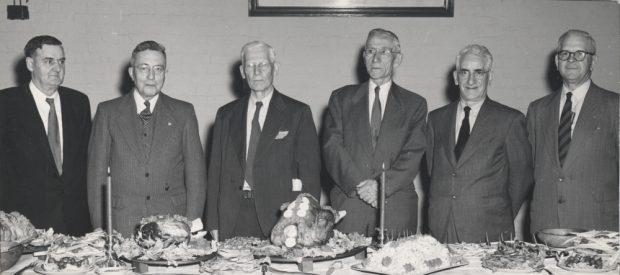 Photographie noir et blanc de six hommes debout derrière une table.    