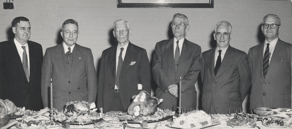 Photographie noir et blanc de six hommes debout derrière une table.    