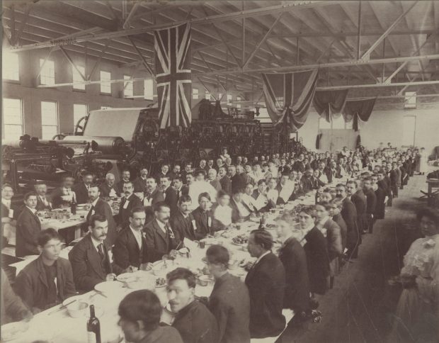 Photographie noir et blanc illustrant plusieurs personnes assises à de longues tables dans une usine. En arrière-plan se trouve une machine à papier. 