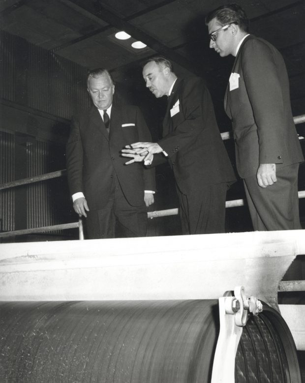 Photographie noir et blanc de trois hommes près d’un grand rouleau. 