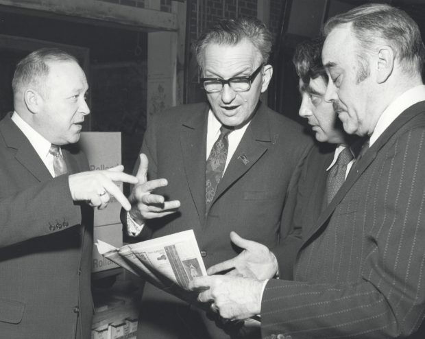 Photographie noir et blanc d’un groupe d’hommes regardant un journal pendant que l’un d’entre eux semble parler. 