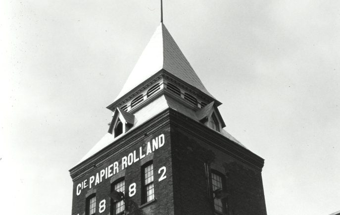 Photographie noir et blanc illustrant une tour reliée à une bâtisse. 