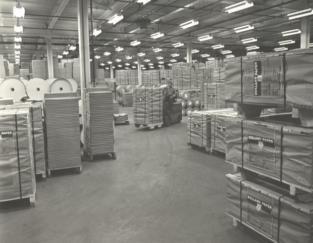 Photographie noir et blanc d’une vaste salle où se trouvent plusieurs emballages et rouleaux empilés.