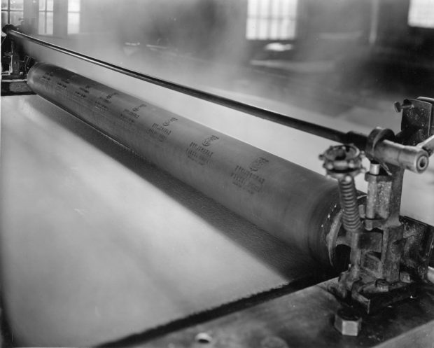 Photographie noir et blanc d’un long rouleau sous lequel passe une feuille de papier. 