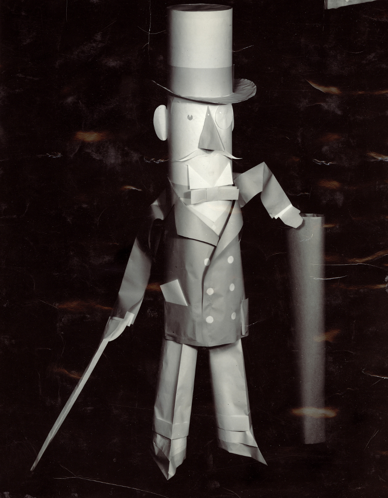 Photographie noir et blanc d’un personnage en habit avec un chapeau haut de forme fait en papier. 