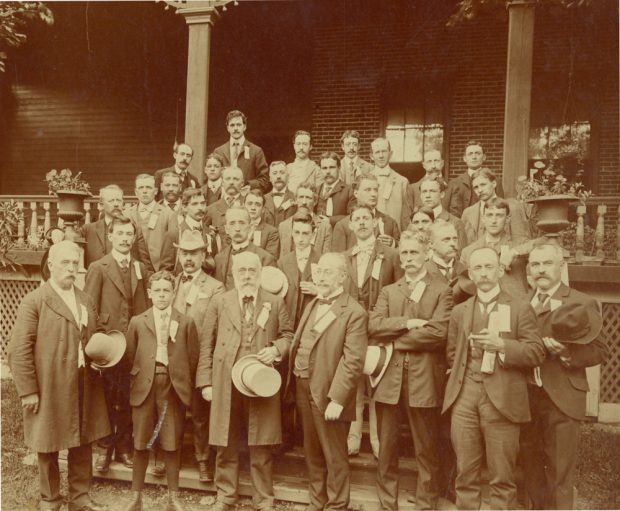 Photographie sépia représentant un groupe d’hommes en complet dans les escaliers devant une maison. 