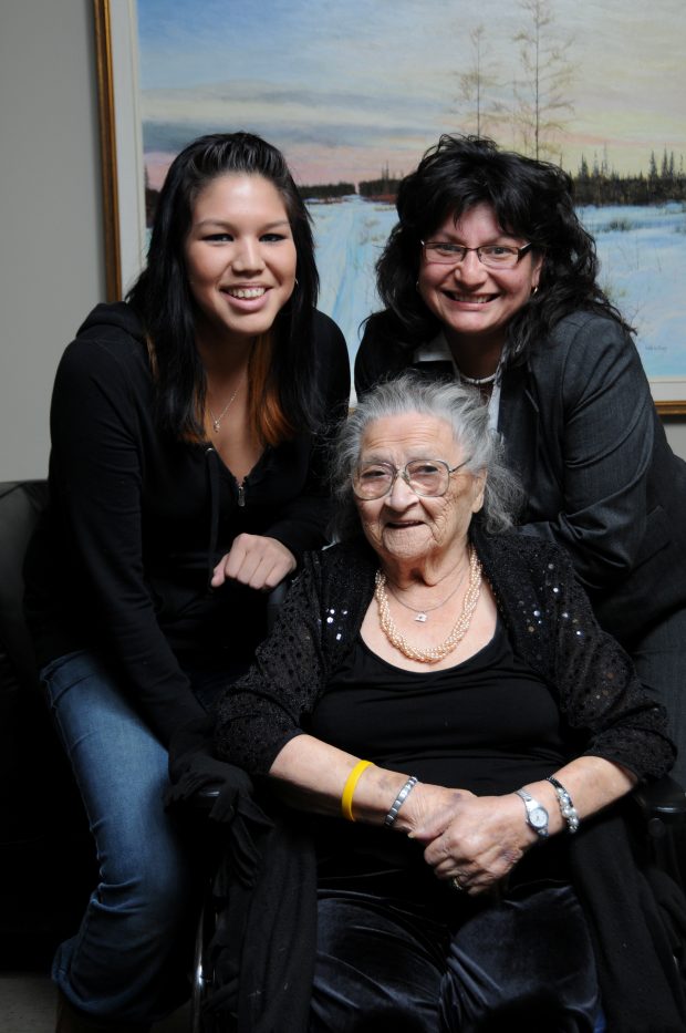 Une femme assise dans un fauteuil roulant avec deux femmes derrière elle. Sur le mur derrière elles se trouve une peinture représentant de la neige et des arbres.