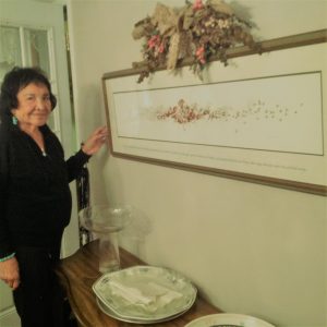 Une Lina Gallup âgée, debout à côté d'une table et d'une photo sur le mur.