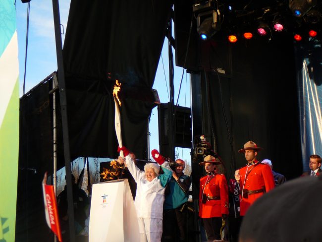 L'aînée Elsie Yanik sur la scène du relais de la flamme olympique en tant que porteuse de la flamme, vêtue d'un authentique survêtement olympique avec des mitaines rouges ornées de feuilles d'érable canadiennes, tenant une flamme blanche. Deux membres de la police montée, en uniforme rouge, se tiennent derrière elle.