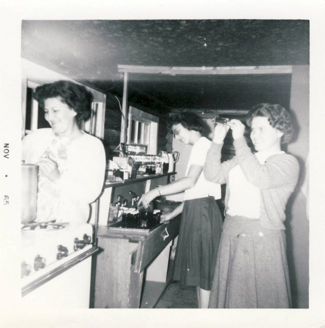 Une Nancy Woodward d'âge moyen se tient dans la cuisine en train de remuer un pot de confiture avec deux autres femmes en arrière-plan, l'une d'entre elles préparant quelque chose sur une autre cuisinière.