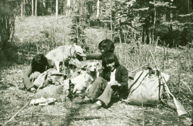 Gerty Sanderson, jeune fille, est assise sur le sol herbeux d'une forêt, à côté d'un grand sac et d'un fusil, avec ses deux jeunes sœurs, à côté de deux chiens, dont l'une caresse l'un d'eux.