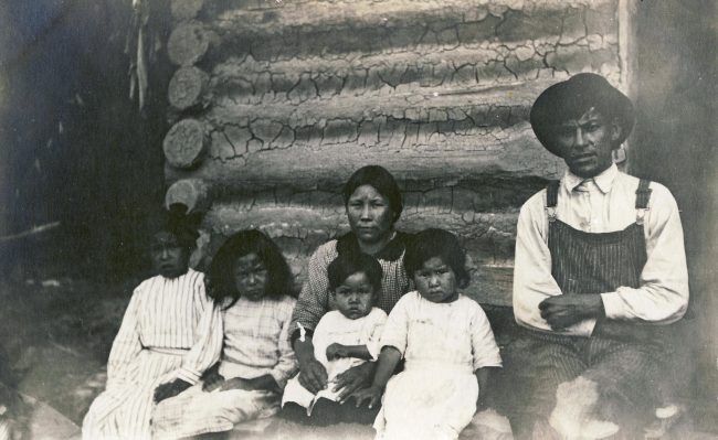 Photo en noir et blanc d'une famille indigène avec quatre enfants assis devant une cabane en rondins.