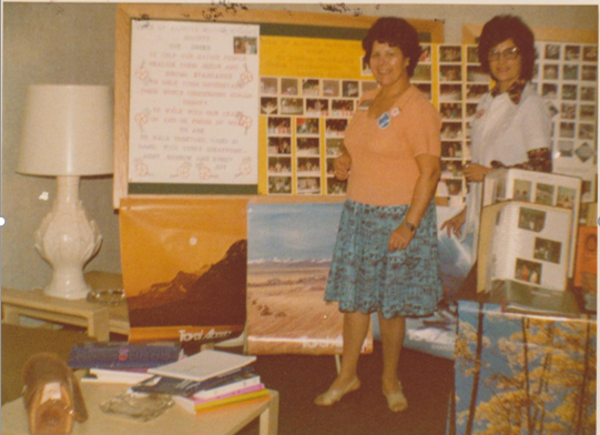 Une photo en couleur de deux femmes entourées par des différents matériaux de présentation, des photos, petites et grosses avec une grande feuille de papier avec de l’écriture. 