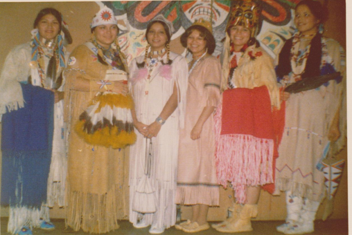 Photo en couleur d'un groupe de jeunes femmes portant des robes traditionnelles et se tenant près les unes des autres sur un fond brun. Les visages des jeunes femmes sont souriants.