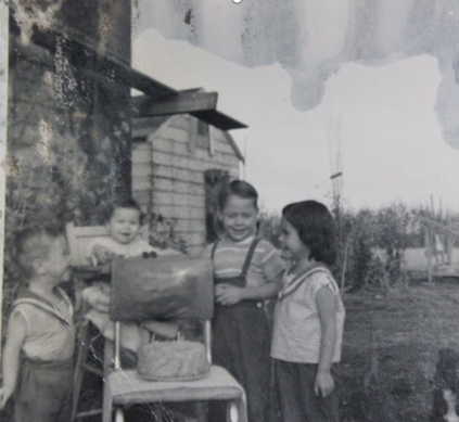 Une photo noire et blanche des jeunes enfants souriants qui se rassemblent autour d’un gâteau brun avec une bougie au dessus. Il y a a côté d’une maison dans l’arrière plan.