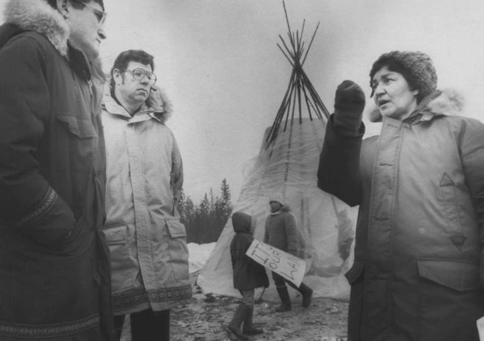 Photo en noir et blanc de deux hommes et d'une femme debout l'un à côté de l'autre, la femme est Dorothy McDonald montrant du doigt et comme si elle parlait aux hommes avec un tipi en arrière-plan et un jeune enfant tenant un panneau de signalisation et quelques arbres au loin.