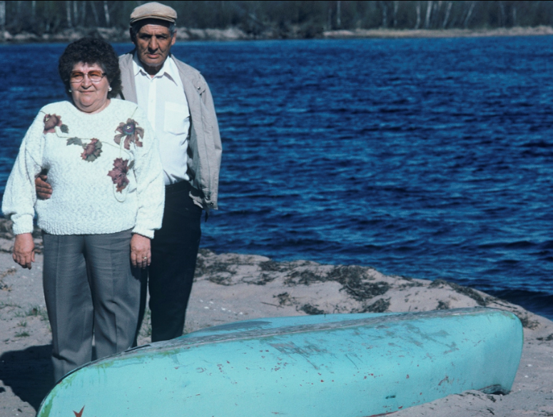 Une vieille dame, Nancy et Bill Woodward, se tiennent à côté d'un canoë bleu clair sur une plage de sable, alors que des vagues sont visibles sur le lac en arrière-plan.