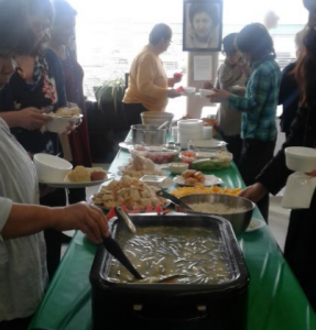 Une photo en couleur d'un groupe de personnes autour d'une table avec les différentes nourritures, la soupe, le bannock, les fruits au long des deux côtés de la table avec une nappe verte en plastique