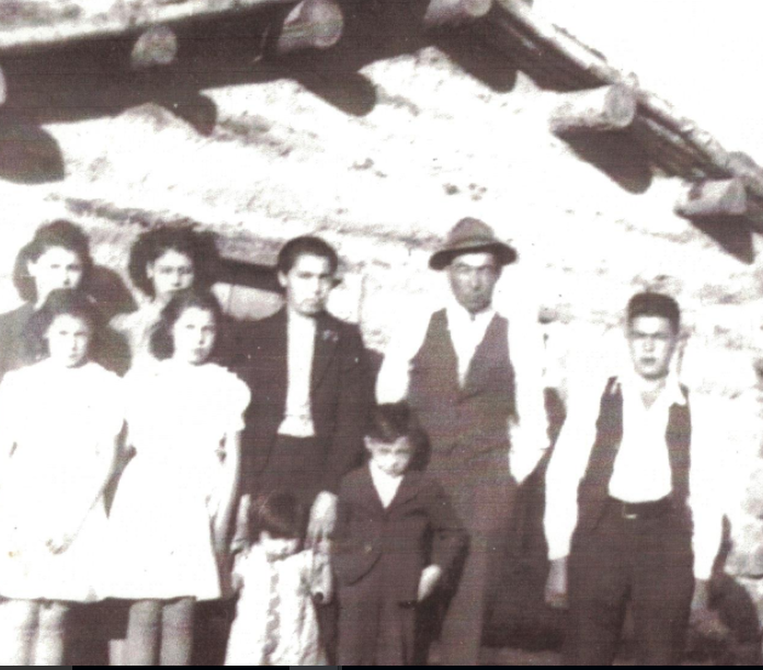 Photo historique du milieu des années 1940 d'une famille dont le père et la mère portent des costumes, entourés d'enfants, devant une maison en rondins.