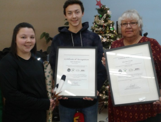 Une jeune femme tenant une plume et un jeune homme et une femme âgée tenant des certificats encadrés regardent la caméra, un arbre de Noël est exposé à l'arrière-plan.