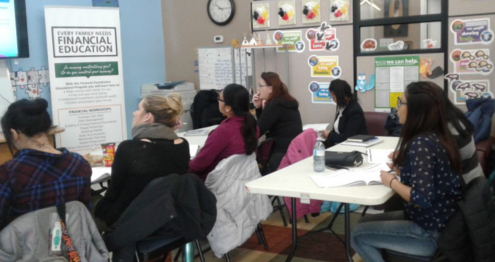 Une photo colorée d'un groupe de femmes qui accessoire devant une table blanche, ils font face en avant. Il y a une affiche qui dit Éducation Financier.