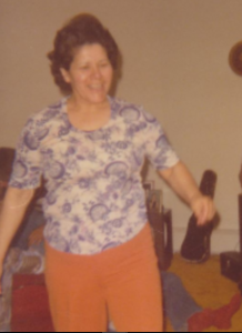 Berta, d'âge moyen, dansant, portant une chemise bleue et blanche à motif cachemire et un pantalon couleur feu.