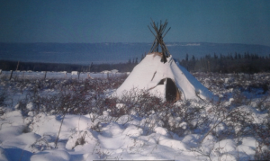 Un tipi recouvert de neige se trouve au milieu d'un champ avec une clôture en fil de fer derrière et une colline boisée en arrière-plan.