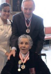 Bertha âgée est assise dans un fauteuil roulant et porte la médaille de l'Ordre du Canada. Le Premier ministre Jean Chrétien se tient derrière elle et une jeune femme se tient à sa gauche.