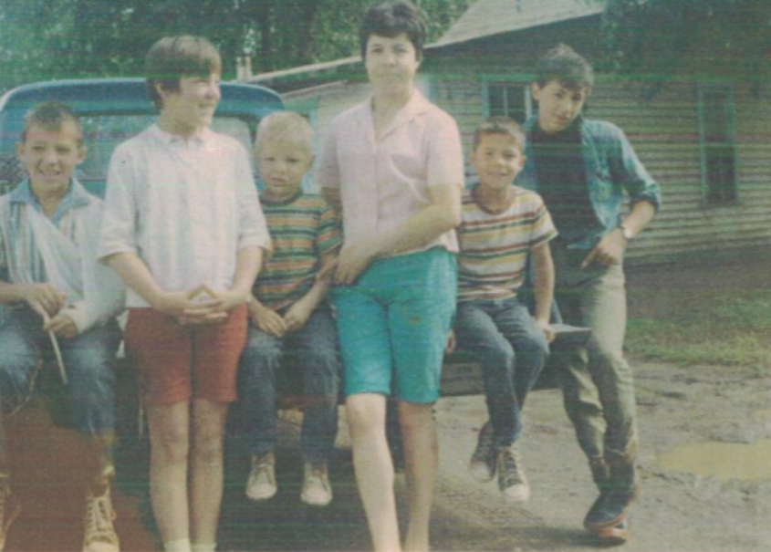 Elsie Cardinal en tant que jeune mère se tenant avec une fille et un fils, trois garçons assis sur le hayon d'un camion bleu dans la cour avant d'une maison.