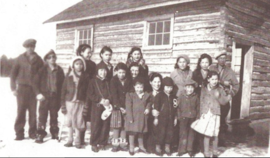 Photo en noir et blanc d'un groupe d'adultes et d'enfants portant des vêtements d'hiver devant un bâtiment en bois avec de la neige au sol.