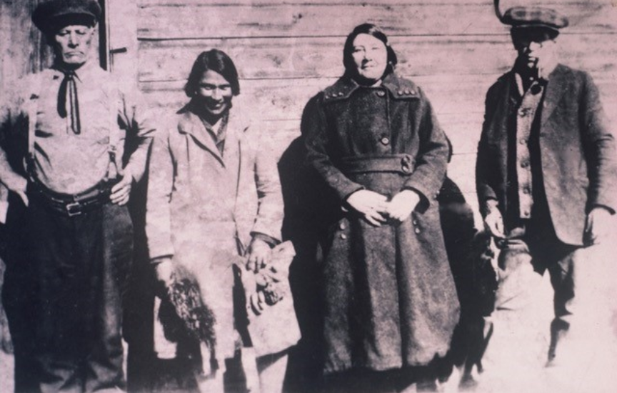Une photo historique de quatre personnes, deux hommes et deux femmes, debout devant une maison en bois, la deuxième personne est la jeune Mary-Rose.