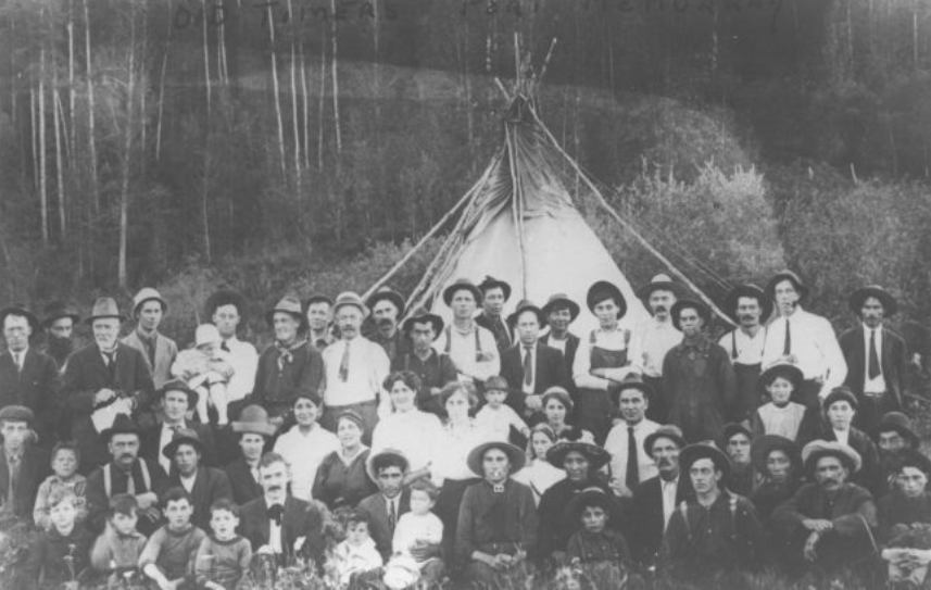 Photo en noir et blanc d'un grand groupe de personnes debout devant un tipi regardant droit devant elles ; certaines sont assises avec de jeunes enfants tandis que d'autres sont debout.