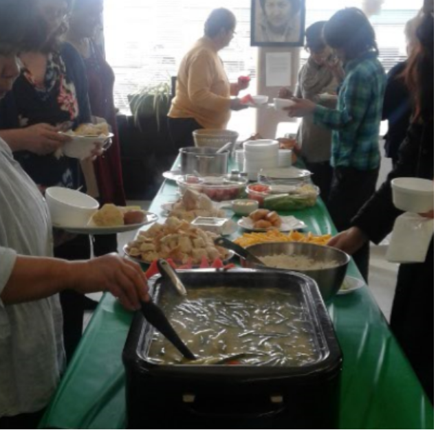 Photo en couleur d'un groupe de personnes autour d'une table avec les différentes nourritures, la soupe, le bannock, les fruits au long des deux côtés de la table avec une nappe verte en plastique