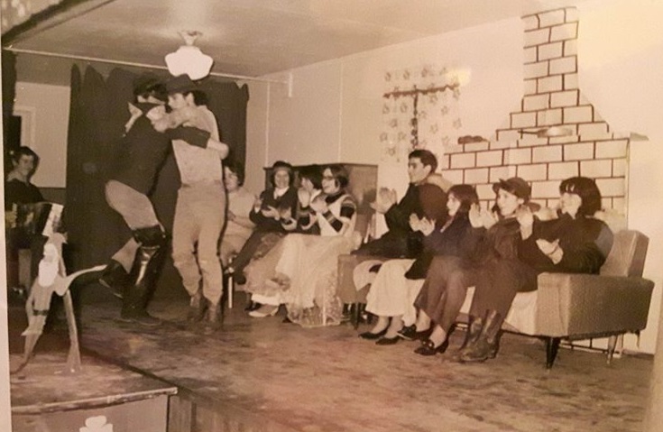 Deux jeunes hommes dansent alors que d’autres jeunes gens assis derrière tapent des mains. Un autre jeune homme joue de l’accordéon.